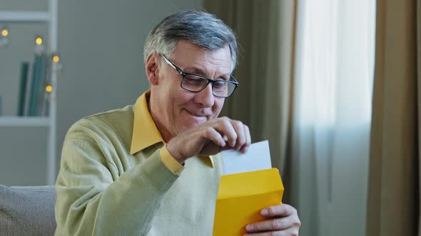 Elderly Caucasian Man Sit at Couch at Home Old 70s Grandfather Holding Paper Letter Reading About