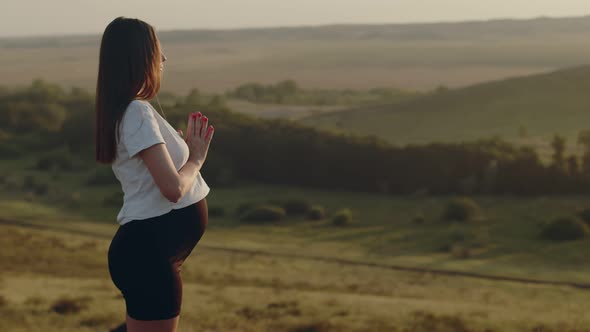 Pregnant Woman Stands Outdoors on Hilltop and Meditates While Doing Yoga Back View