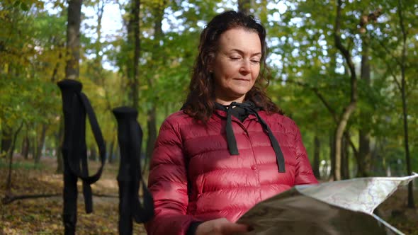 Portrait of Thoughtful Caucasian Mature Woman Examining Paper Map Standing in Autumn Park with Poles