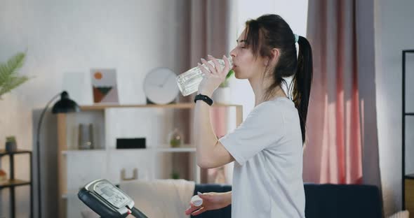 Woman with Ponytail Drinking Water During Training on Treadmill at Home