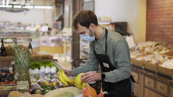 Male Assistant in Supermarket Food Store Worker in Medical Mask and Apron Arranges Bananas on