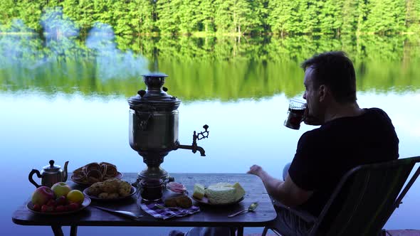Man drinking tea by the lake. Vintage tea samovar with smoke and food near lake