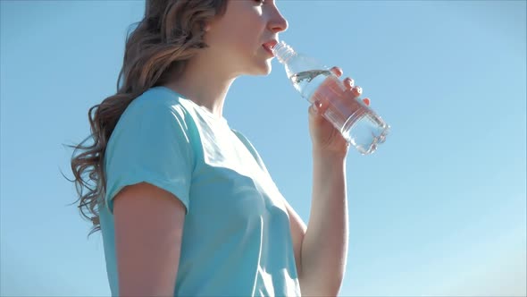 Woman of European Appearance in the Open Air Drinks Water From a Bottle Against a Clear Blue Sky