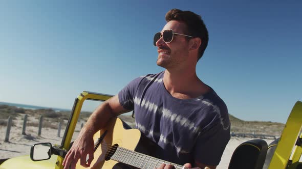 Happy caucasian man sitting in beach buggy by the sea playin gguitar