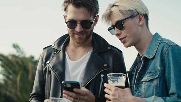 Two young caucasian men browsing phone and having fun on music festival while drinking beer. Shot wi