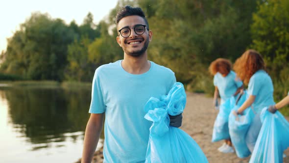 Young Arab Volunteer with Trash Bag Smiling on Lake Shore with People in Background
