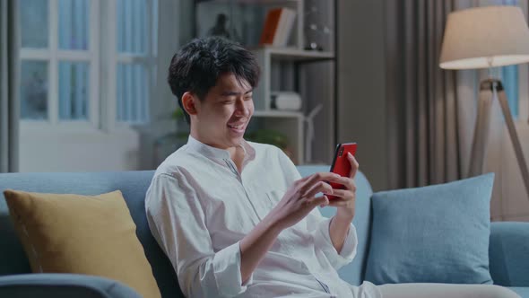 Asian Man Touching Smartphone And Laughing While Sitting On Sofa In The Living Room