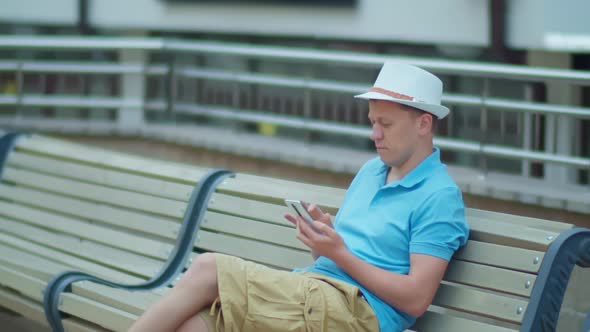 Man in hat uses mobile phone while sitting on city bench, camera tracking