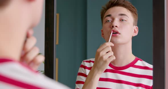 Young gay man queer holding lipstick or lip gloss and paints lips and doing make up
