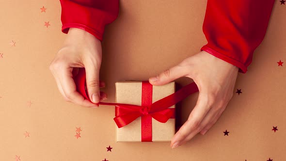 Human hands with red sleeves untied a brown paper gifts with a red satin ribbon bow on brown back alt