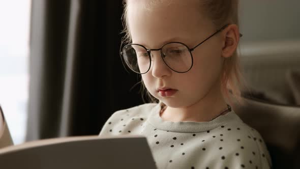 Little child clever girl reading a book in glasses after school