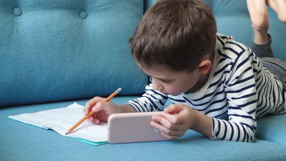 A Child in a Room Lying on a Sofa Uses the Internet on a Smartphone for Remote Learning