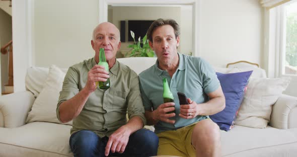 Portrait of happy caucasian grandfather with adult son with beer watching tv in living room