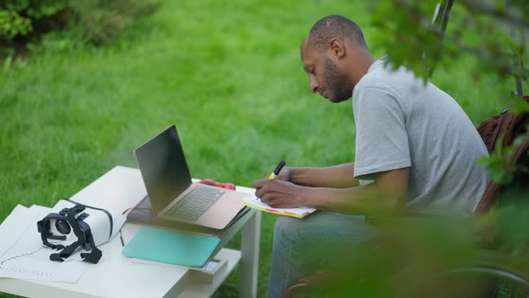 Side View of Concentrated African American Man Writing in Sketchpad Sitting on Spring Backyard with