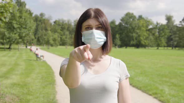 A Young Caucasian Woman in a Face Mask Points at the Camera and Nods on a Pathway in a Park