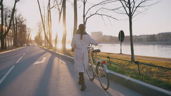 Woman in Beige Coat Carries Yellow Bicycle Along Road