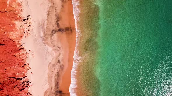 Red cliffs lining sandy beach as small waves break on sand, straight down aerial