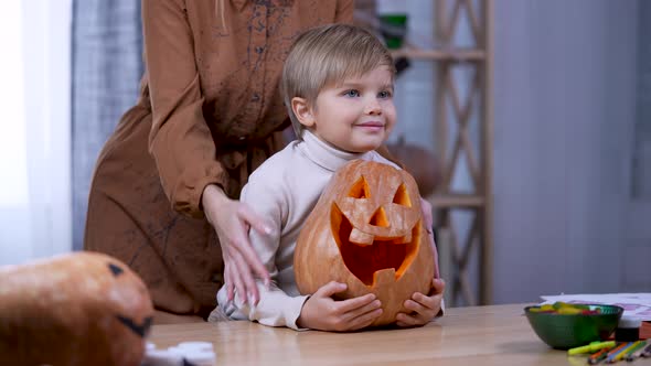 The Boy Sits at a Table Looks Ahead and Hugs a Large Orange Pumpkin