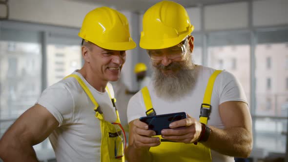 Two Builders in Hardhat and Overall Using Smartphone at Construction Site