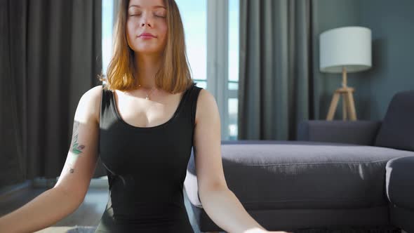 Young Caucasian Woman in Black Jumpsuit Sitting in Lotus Position and Meditating Yoga at Home