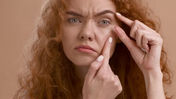 Concerned RedHaired Young Woman Searching Eye Wrinkles Over Beige Background
