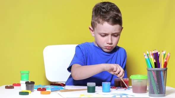 A little preschooler boy is sitting at a table, drawing with watercolor paints on a sheet of paper.