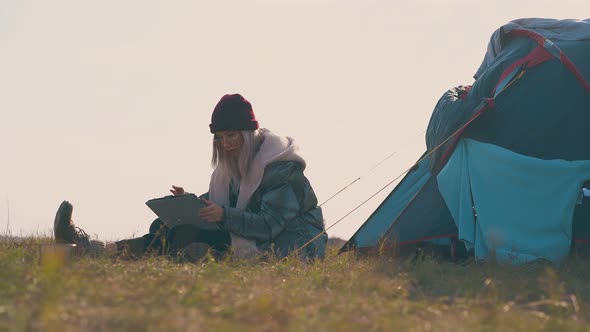 Woman Hiker with Tablet Sits at Tent on Autumn Meadow