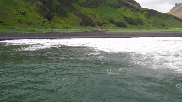 Waves crashing on black sand beach in Vik, Iceland with drone over water moving forward.