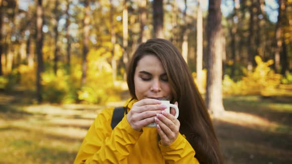 Young Woman in Parka Coat and Backpack is Holding Mug of Tea Inhaling Its Aroma and Smiling Walk in