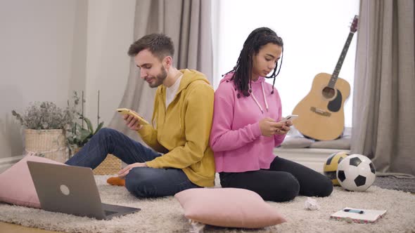 Young Multiethnic Couple Sitting Back To Back on the Floor and Using Smartphones. Girlfriend Spying