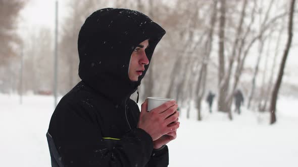 Young Guy Winter After Running in Park with Red Nose in Black Jacket Stands and Holds Mug with Hot