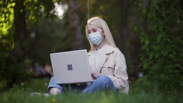 A Blonde Woman in a Medical Mask is Talking By Video Call Using a Laptop in the Park