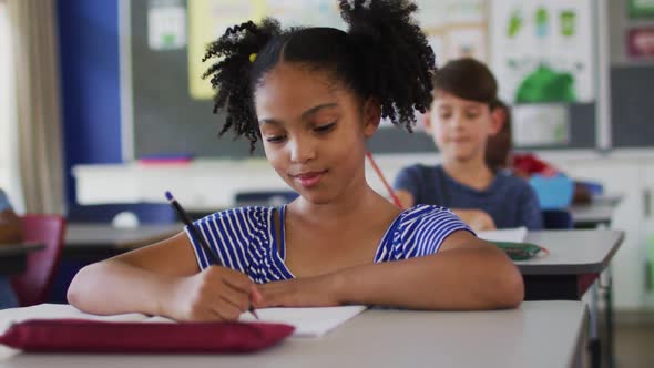 Portrait of happy mixed race schoolgirl sitting at classroom, making notes, looking at camera