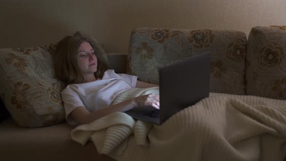 Young Girl Lying on a Sofa with a Laptop and Typing Message, Night Time