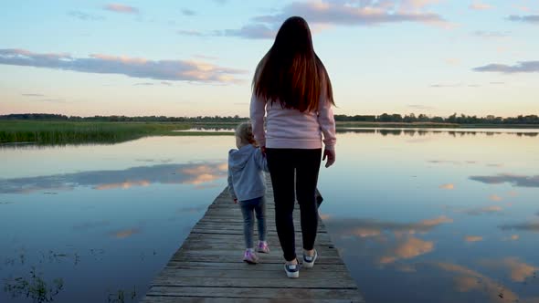 Mom and Little Daughter Walk on the Bridge at Sunset. There's a Lake Around the Bridge