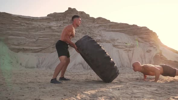 Two Athletes Train in Active Mode on the Beach Doing push-UPS and Pushing a Huge Wheel