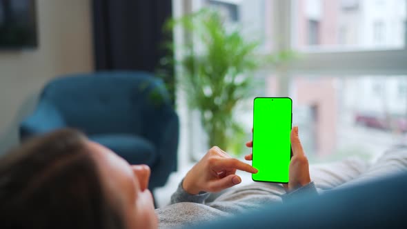 Woman Lies at Home on the Sofa and Using Smartphone with Green Mockup Screen in Vertical Mode