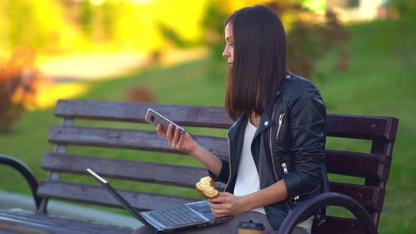 Attractive Young Girl Eating a Bun and Working on a Laptop, Talking on the Phone