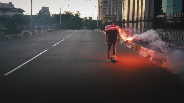 Young Man in Flag of USA Tied on His Neck is Riding Skateboard Along Deserted Street with Burning