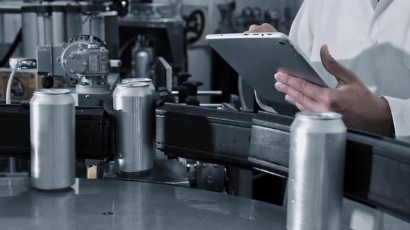 Male Worker Types on a Tablet Near Conveyor with Cans at Beverage Factory Production Line Close Up