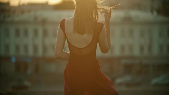 A Young Graceful Woman in Red Dress Standing on the Roof and Fixing Her Hair - Sunset