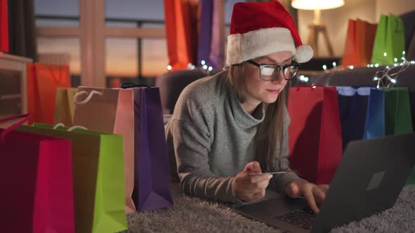 Woman with Glasses Wearing a Santa Claus Hat is Lying on the Carpet and Makes an Online Purchase