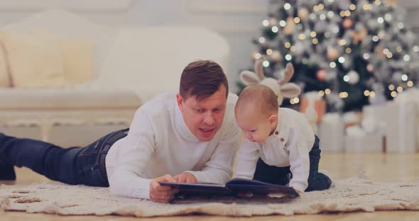 Happy Dad with His Son Lie By the Beautiful New Year Tree and Read a Book with Fairy Tales