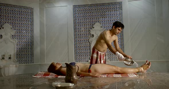 Woman Is Lying on Warm Marble Table in Hammam and Male Massagist Is Pouring Her Body By Water