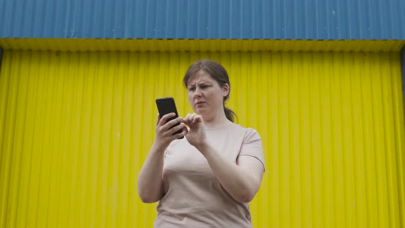 Woman Clicks on Smartphone Screen is Very Surprised and Upset Near Yellow Wall