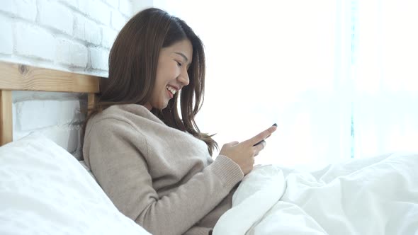 Asian woman in bed checking social apps with smartphone.