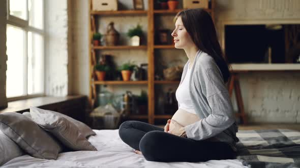 Cute Pregnant Girl Is Stroking Her Belly Resting on Bed in Modern Flat and Looking Out of the Window