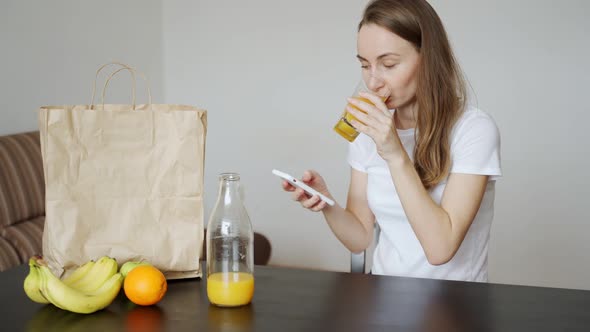 Woman Drinks Orange Juice and Uses a Mobile Phone While Relaxing in the Kitchen at Home