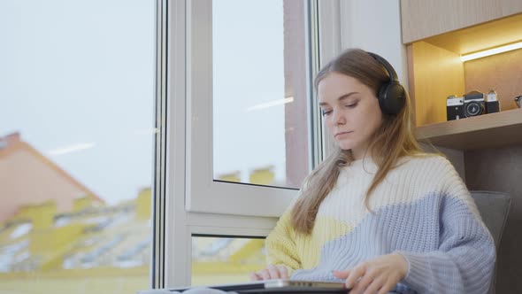 A young woman at home by the window puts on headphones and opens a laptop and gets ready for work.