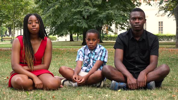 A Black Family Sits on Grass in a Park and Looks Seriously at the Camera